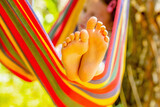 Fototapeta Natura - Young girl relaxing in hammock while working on laptop during camping day in nature. She communicates online, learns remotely on a background of green lawn. Selectve focus on bare feet