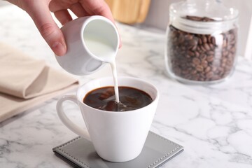 Wall Mural - Woman pouring milk into cup with aromatic coffee at white marble table, closeup