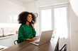© Mia_Bon - A joyful young woman with afro hairstyle is on her phone calling while working with laptop at her home office with water beside her