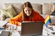 © LIGHTFIELD STUDIOS - Joyful teenage girl looking at laptop and enjoying e-learning next to headphones and pride flag