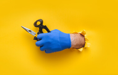 Wall Mural - A left man's hand in a blue fabric knitted glove holds wire cutters and pliers. Torn hole in yellow paper. The concept of a worker, a labor migrant, a master of his craft. Copy space.