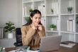 © wichayada - Young Asian business woman sits and works using a laptop in a modern office decorated with shady green plants.
