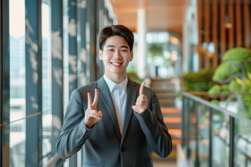 Wall Mural - Portrait of a successful smiling Asian male businessman standing in a suit in an office center and showing ,the super sign with his fingers to the camera, Generative AI