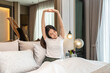 Asian woman is smiling and raising arms to stretching on the bed in bedroom with feeling refreshed and relaxed