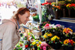 Young adult joyful woman kneels to choose buy colorful potted flowers at outdoor market stall. Sale of blooming plants in pots at botanical street German store on sunny spring day