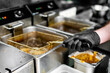 © pavel siamionov - person in black gloves is frying food in professional kitchen using deep fryer. The stainless steel fryer contains bubbling oil, and adjacent containers suggest a busy commercial kitchen environment.