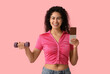 Happy young African-American woman with sweet chocolate bar and dumbbell on pink background