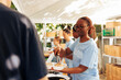 © DC Studio - African american woman serving free food to the poor, needy individual at an outdoor food bank. Female volunteers feeding and providing support to the hungry and less privileged at a homeless shelter.