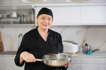 Wall Mural - Positive adult woman in cook uniform holding frying pan in kitchen