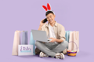 Sticker - Young Asian man in bunny ears with mobile phone and laptop shopping online on lilac background. Easter Sale