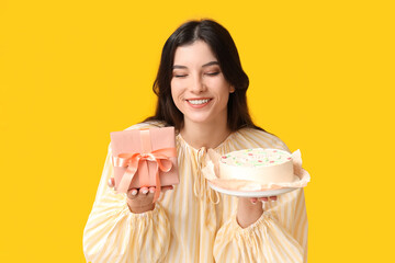 Poster - Happy young woman with sweet bento cake and gift box on yellow background. International Women's Day