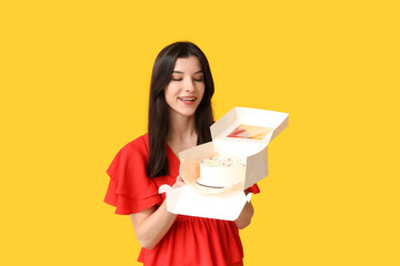 Poster - Beautiful young woman holding box with sweet bento cake on yellow background. International Women's Day