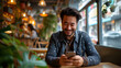 Cheerful man enjoying his time, smiling while looking at his smartphone in a lively, plant-filled coffee shop setting.
