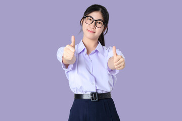 Wall Mural - Portrait of Happy young Asian student girl in school uniform shows thumbs up isolated on purple background.