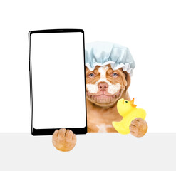 Wall Mural - Smiling Mastiff puppy wearing shower cap, with cream on it face holds rubber duck and big smartphone with white blank screen in it paw. isolated on white background