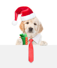 Sticker - Cute Golden retriever puppy wearing necktie and santa hat holds gift box and looks above empty white banner. isolated on white background