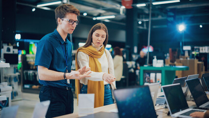 Young Woman Having a Conversation with a Salesperson in Home Electronics Store while Selecting a Laptop. Stylish Customer Wishes to Buy a Computer Device. Shopper Explores Latest Options with Expert