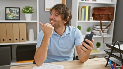 Wall Mural - Cheerful young caucasian man confidently flashing a thumbs-up, joy radiating from his open-mouth smile, while pointing at his smartphone in the office background