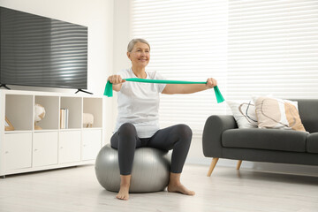 Wall Mural - Senior woman doing exercise with elastic resistance band on fitness ball at home