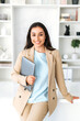 © Kateryna - Vertical photo of successful arabian or indian young business woman in a suit, digital manager, seo, holding laptop while standing in a modern office space near work desk, looks and smile at camera