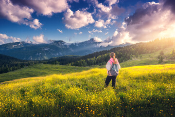 Wall Mural - Girl on the hill with yellow flowers and green grass in beautiful alpine mountain valley at sunset in summer. Landscape with young woman in alps, trees, sky with clouds. Hiking. Spring in Slovenia