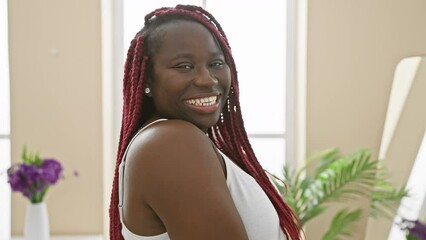 Canvas Print - African american woman with red braids smiling in a bright living room with plants.