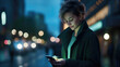 Young woman in a green jacket is focused on her smartphone at night with city lights creating a bokeh effect in the background.