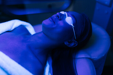 Top view of female client having blue LED light facial photodynamic therapy treatment in beauty salon. Young woman lying during non-invasive type of phototherapy. Concept of beauty treatment.