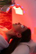 © dikushin - Vertical shot of young woman having red LED light facial photodynamic therapy treatment in beauty salon. Luxury medical clinic using phototherapy device with red light wavelength for acne treatments.