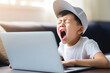 yawning 10 yo Asian boy in white t-shirt and baseball cap sitting in front of an open laptop. themes of boredom, tiredness, online learning, remote work, youth culture, childhood, and technology