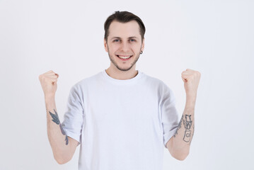 Wall Mural - Attractive caucasian young man with tattoos in casual clothes looking at camera, smiling and showing winner gesture isolated on white studio background