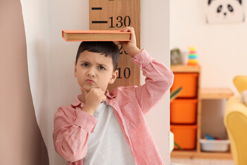 Sticker - Thoughtful little boy measuring height with book near wooden stadiometer at home