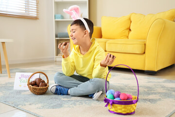 Poster - Cute little boy in bunny ears headband eating chocolate eggs for Easter at home