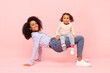 © Home-stock - Joyful little black daughter having fun with young mother, girl sitting on mom's legs like on chair against pink studio background