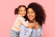 © Home-stock - Happy black mother posing with her cute little daughter, girl hugging mom from back and smiling together at camera, pink studio background