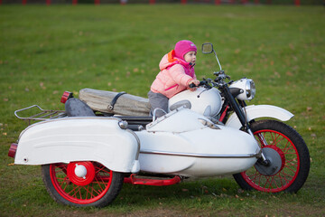 A little girl is sitting on a white vintage sidecar three wheel motorcycle isolated on green grass background. Horizontal image outdoors. 