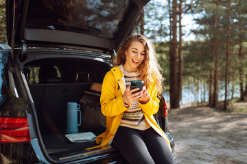 Wall Mural - Young woman sitting in open trunk with phone. Lifestyle, travel, tourism, nature, active life.