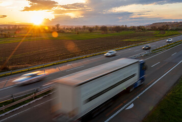 Wall Mural - A big truck and several cars  drive at high speed on the highway through the rural landscape. Fast blurred highway driving. A scene of speeding on the highway. Beautiful sunset in the background.