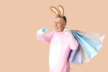 Wall Mural - Senior man in Easter bunny costume with shopping bags and credit card on beige background