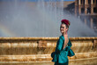 Young, pretty, blonde woman in typical green colored flamenco suit, posing with her back next to a fountain with rainbow reflected in the water. Flamenco concept, typical Spanish, Seville, Andalusia.