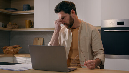 Wall Mural - Caucasian busy freelancer man working distant from home work with laptop at kitchen exhausted businessman take off glasses suffering from eye strain pain job break from computer dry eyes overworking