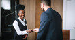 © ihorvsn - Rich good-looking male customer checking into luxury hotel and handing over passport to welcoming female African receptionist. Handsome busy manager talking on phone on background.