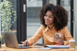 © Songsak C - Portrait of a beautiful confident businesswoman using a laptop computer holding a mobile phone sitting in a modern office. Smiling African American freelancer working online from home.