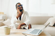 © SHOTPRIME STUDIO - Smiling African American Man Working on Laptop in Cozy Home Office In this image, an African American man is sitting comfortably on a sofa in his living room, wearing a bathrobe and glasses He is a