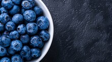  a close up of a bowl of blueberries on a black surface with drops of water on the top of the bowl.