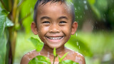 Fototapeta  - A happy young boy with a beaming smile enjoying a refreshing rain shower among green leaves.
