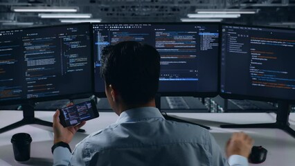 Back View Of Asian Man Developer Working With Mobile Phone While Write Code With Multiple Computer Screens In Data Center