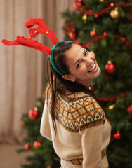 Poster - Portrait, happy and woman in home for Christmas, party or holiday in apartment in Spain. Xmas, face and smile of female person in antlers for festive celebration, funny and laughing by tree in house