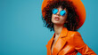 Portrait of a glamorous female model in sunglasses, a hat and a bright orange leather suit, with curly hair on a blue background in a gradient style, with copy space for text. Style and high fashion