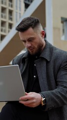 Sticker - Young man with a laptop on fresh air. Handsome young man in a coat sitting in the street and working on a modern device in the city. Distance work. Vertical video.